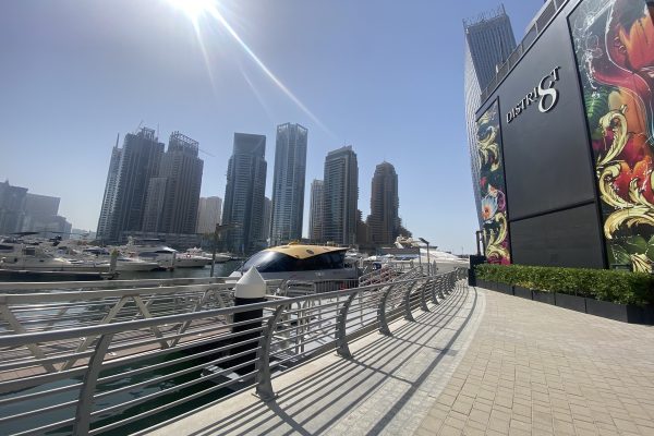 Boat Tour in Dubai: A bright, sunny view of the Dubai Marina waterfront, showing a pier, docked yachts, the District 8 building with a colorful mural, and a row of tall skyscrapers in the background.